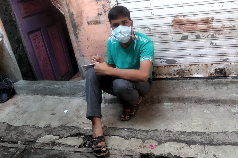 A teenaged boy dressed in blue jeans and a blue shirt sits on a stoop writing in a notebook. He is wearing a protective face mask.