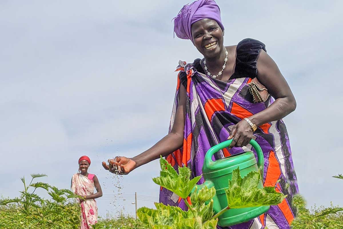In South Sudan, a woman smiles broadly while watering her thriving garden. Another women smiles from the background.