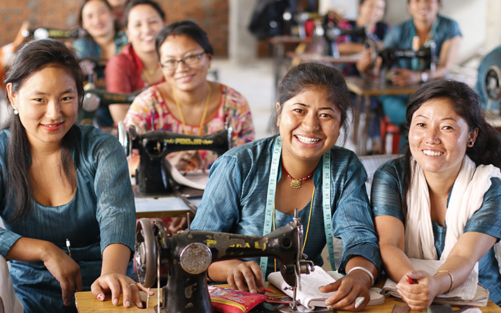 Three women sitting behind a sewing machine smile toward the camera.