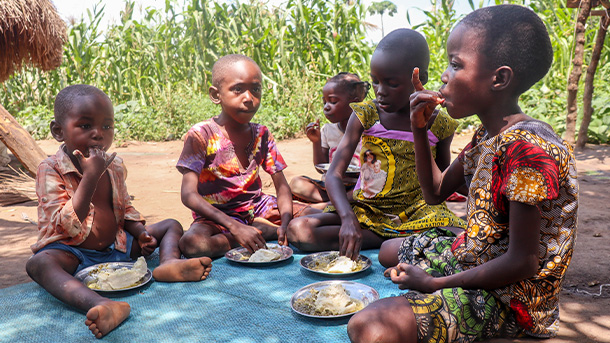 Five children are outside seated on a blanket sharing food from four plates.