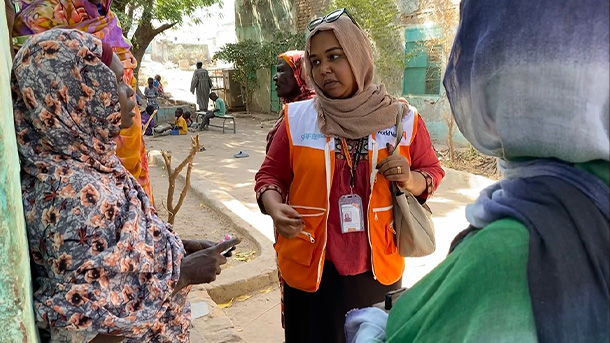 A woman stands outside wearing a humanitarian vest with the World Vision logo while speaking with a few women.