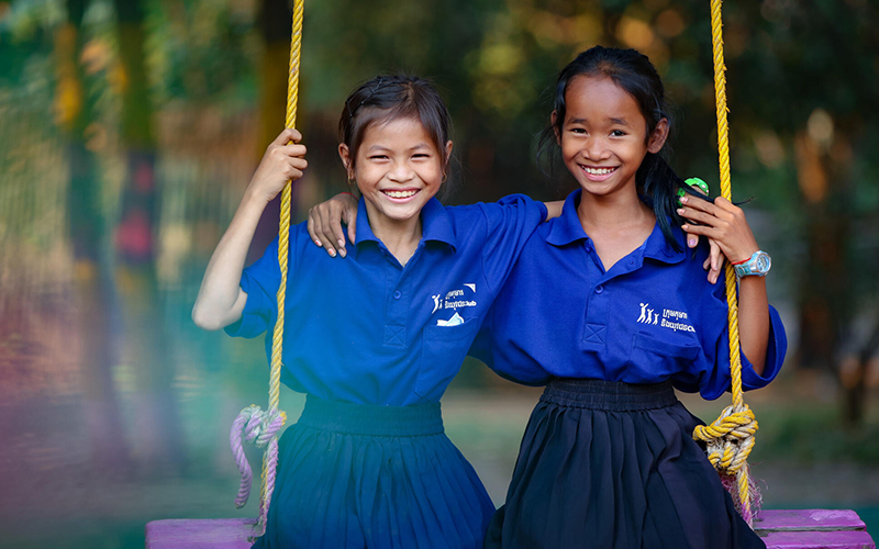 Two young girls dressed in school uniforms sit side by side on a purple swing, embracing each other and smiling.