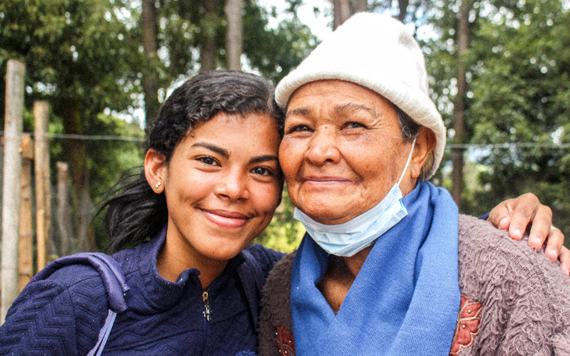 A teenage girl and older woman outside smile and embrace against a backdrop of tall green trees.