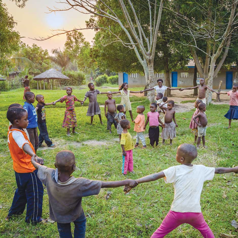 A group of children hold hands and form a circle around another group of children as they play in the grass.