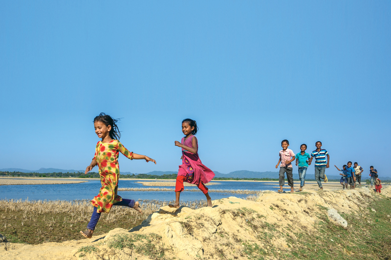 Against a clear blue sky, a group of small children form a line as they run along a shoreline.