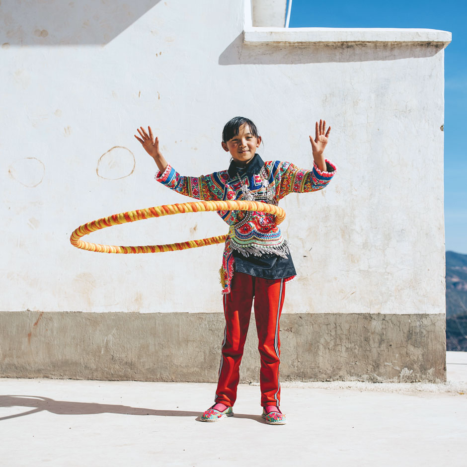 A young girl in colourful clothing hula hoops in front of a white concrete wall.