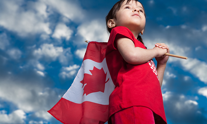 young child holds a Canadian flag while looking up at a blue sky.