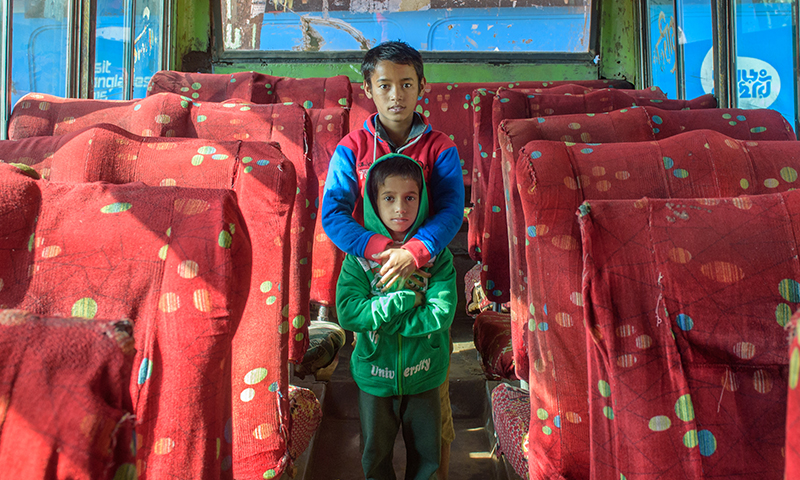 an older boy stands behind a younger child with arms protecting, while standing inside a bus