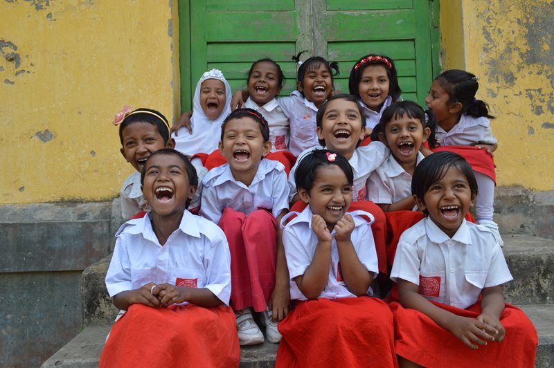 A group of 8-year-old girls from Bangladesh laugh as they sit on steps in front of their school. They are wearing their school uniform which consists of a white, short sleeved, button-down shirt and orange skirts.