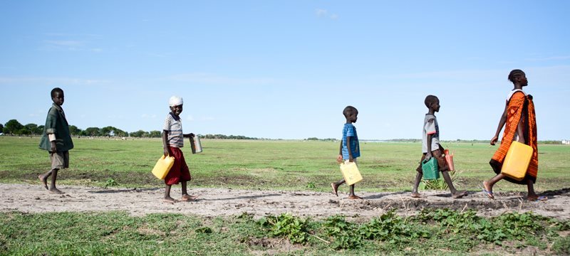Two children walking through a field, one carring a jug for water, smile at the camera