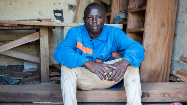 A young man from DRC sits on a pile of timber in a carpentry workshop.