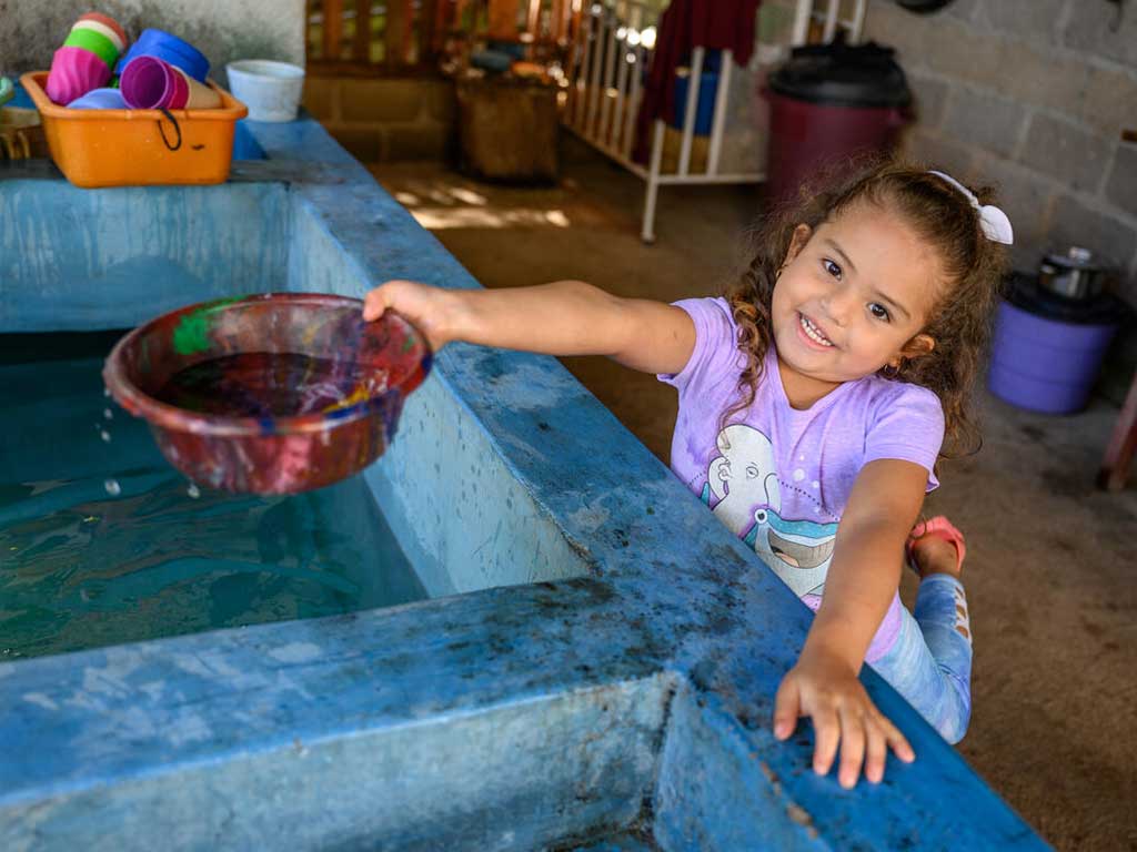In Honduras, a little girl with a white headband is smiling at the camera and dipping a red bowl into a basin of clean water.