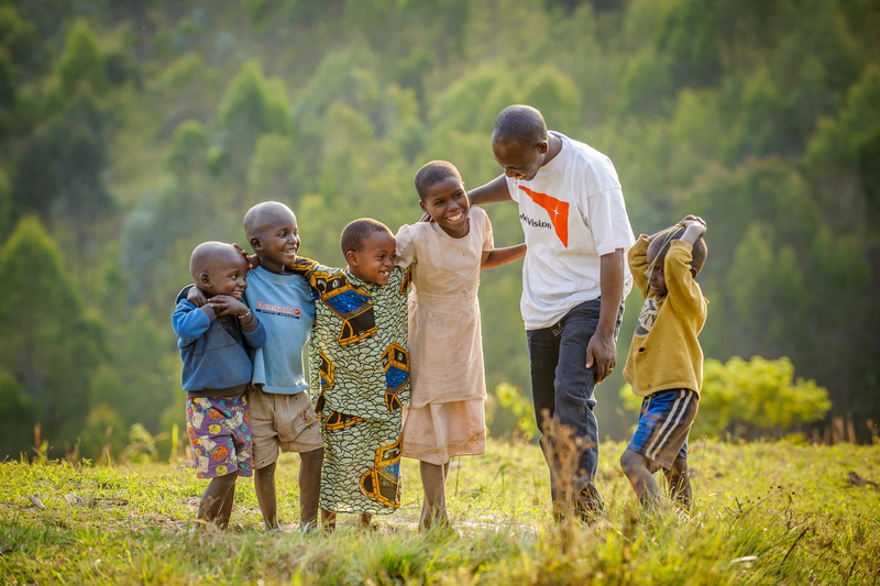 A World Vision staff member is joined by five children of various ages in a grassy field on a sunny day.