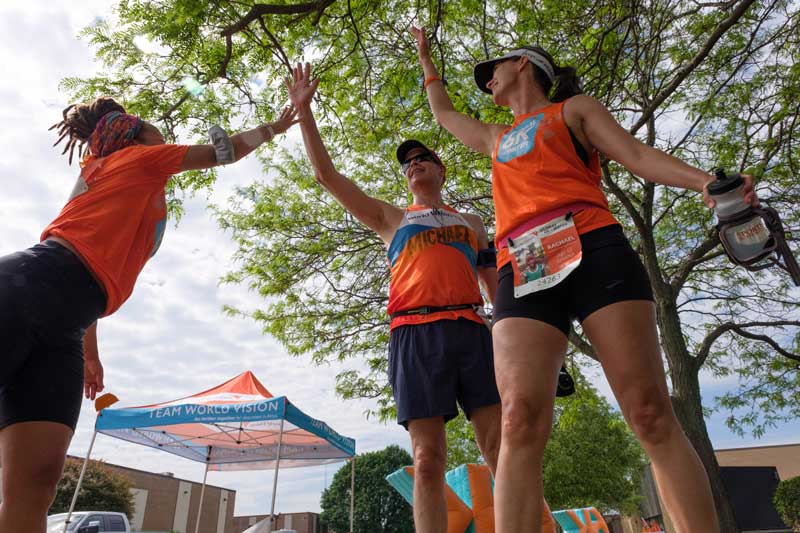 Three adults stand outdoors under a tree. Each is wearing an orange racing bib and black shorts. They raise their hands together for a group high-five.