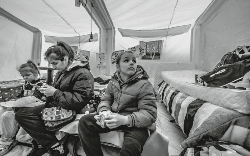 A black and white image shows a young girl looking up in a tent, with a woman and  younger child beside her.