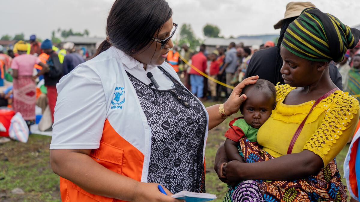 A woman wearing an orange and white vest with the World Food Programme logo on it places her hand on an infant's head, talking to the baby's mother.