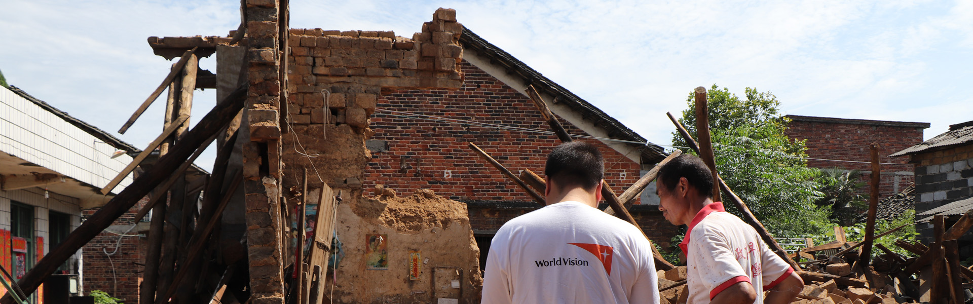 A man wearing a white t-shirt with a World Vision logo faces a building that has been reduced to rubble.