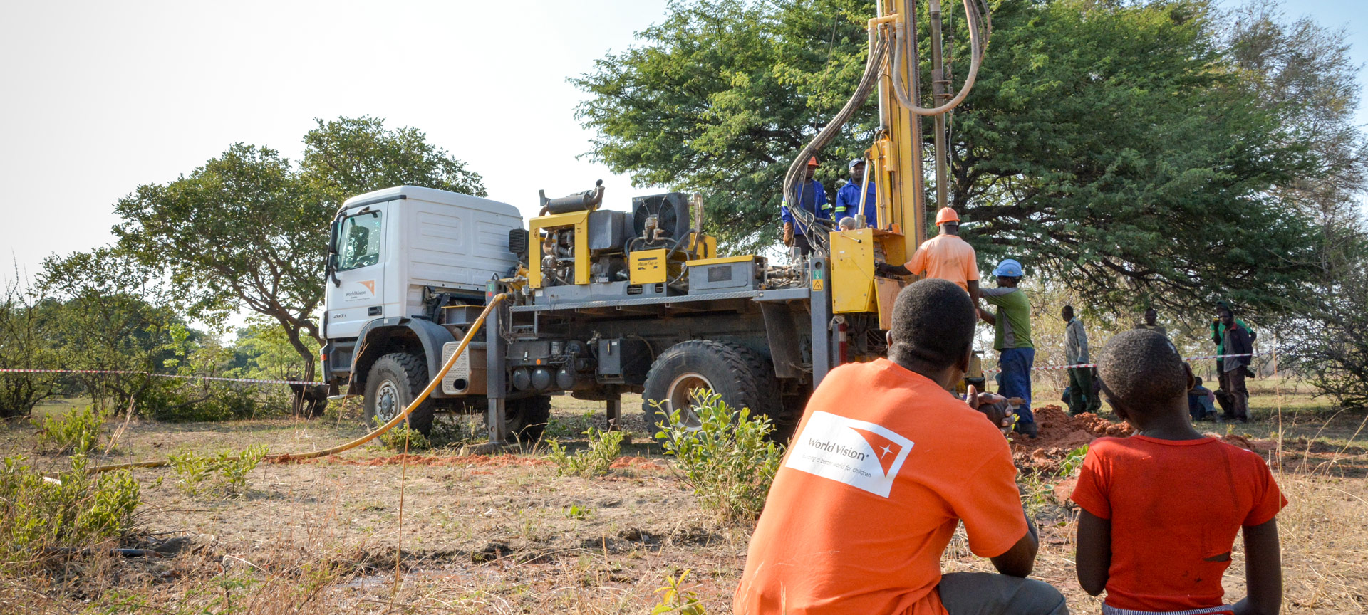 A boy stands beside a man in an orange World Vision shirt, watching a well-drilling rig. 