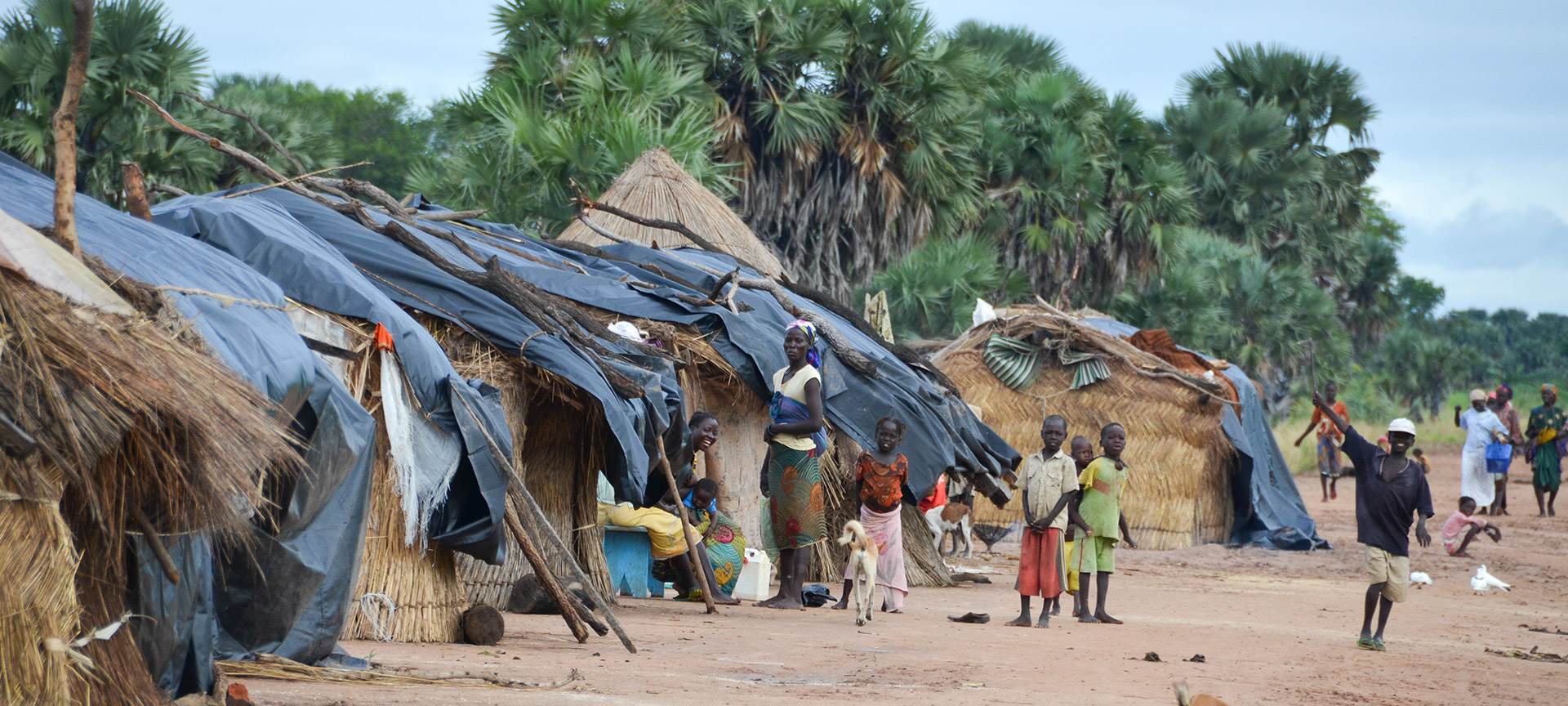 Women and children dressed in colorful clothing gather outside a line of small cabins made of straw. 