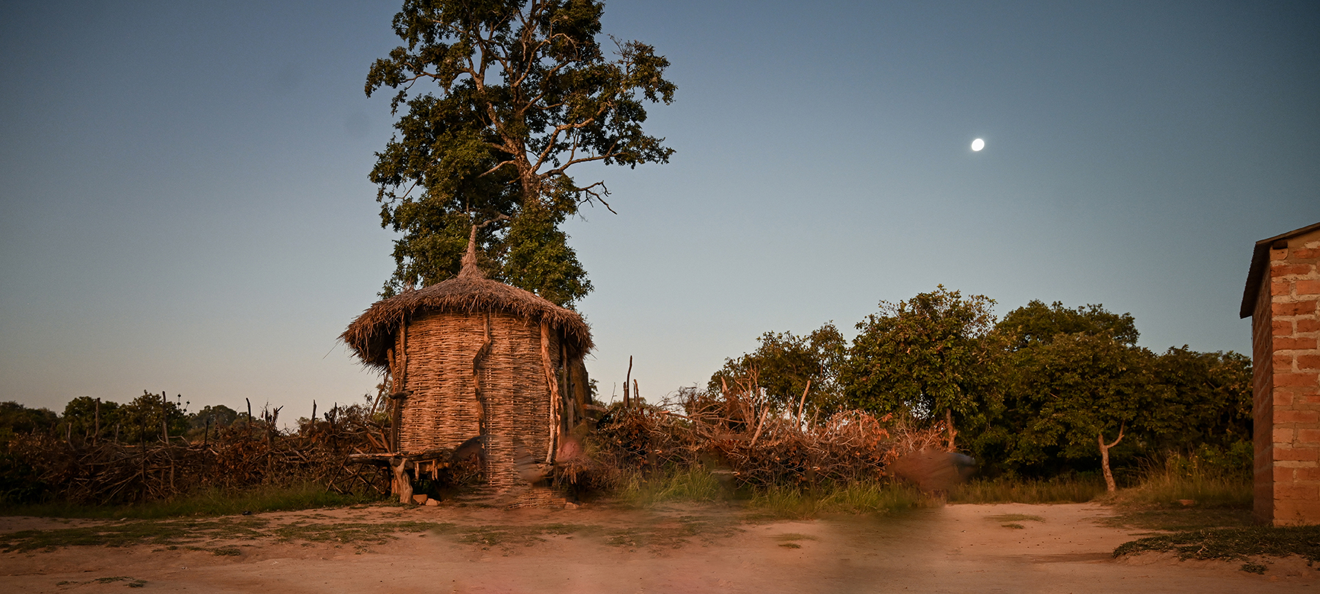The sunsets over a rural area where a small cabin made of straw and a brick building stand. 