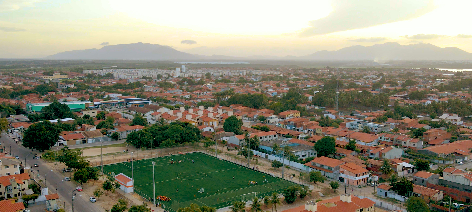 A birds-eye view of the sun rising over a town. Homes with orange roofs fill the town and a green football field can be seen. 