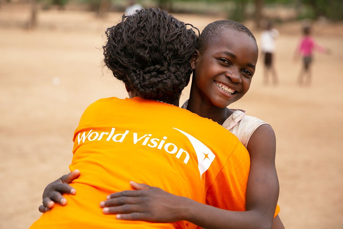 A World Vision staff member in an orange shirt hugs a smiling child.