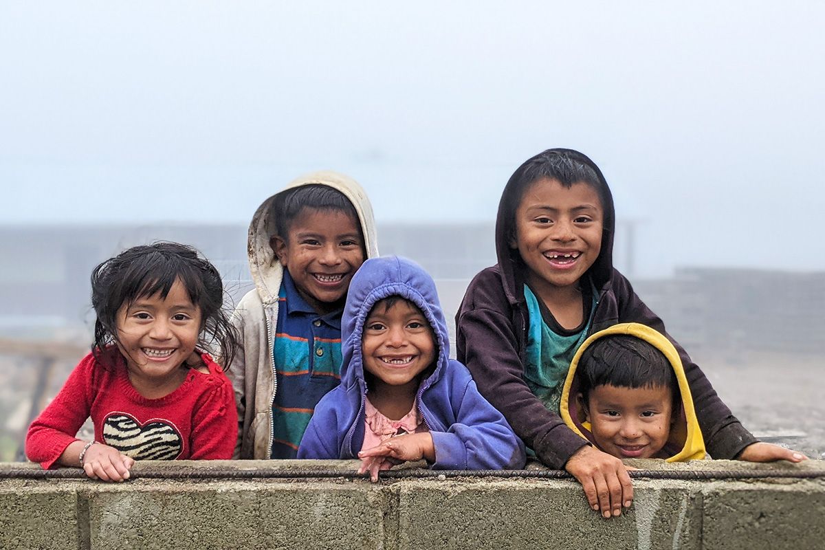A group of four children wearing colourful sweaters, standing outside behind a ledge smiling.