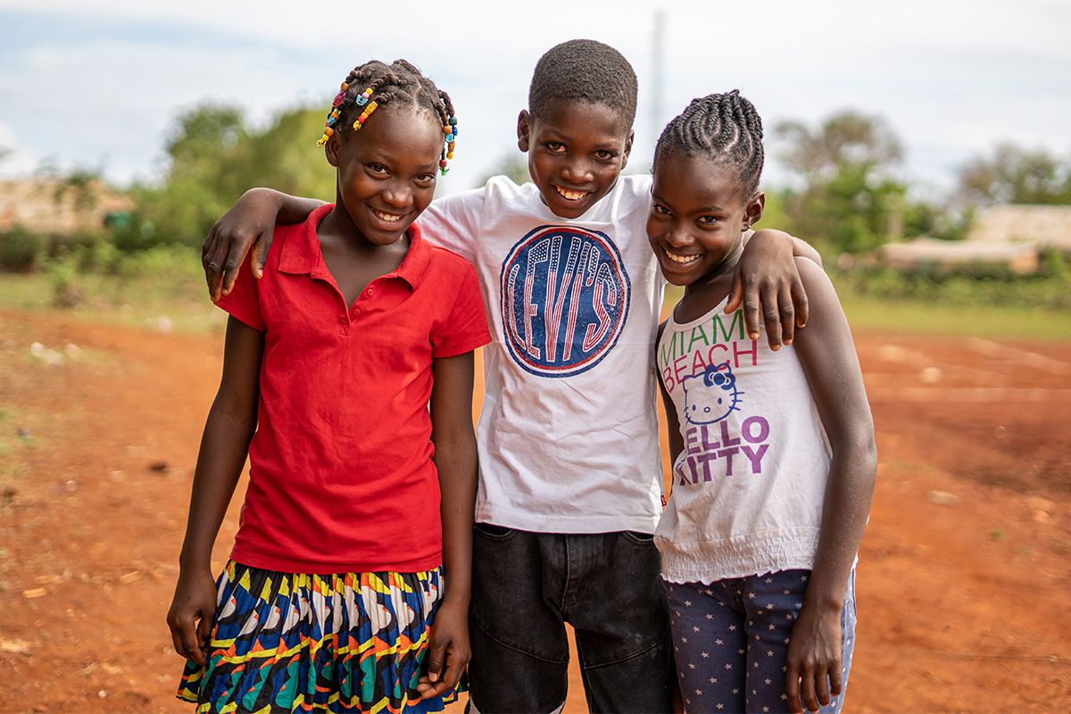 A group of three children standing outside smiling and embracing each other. 