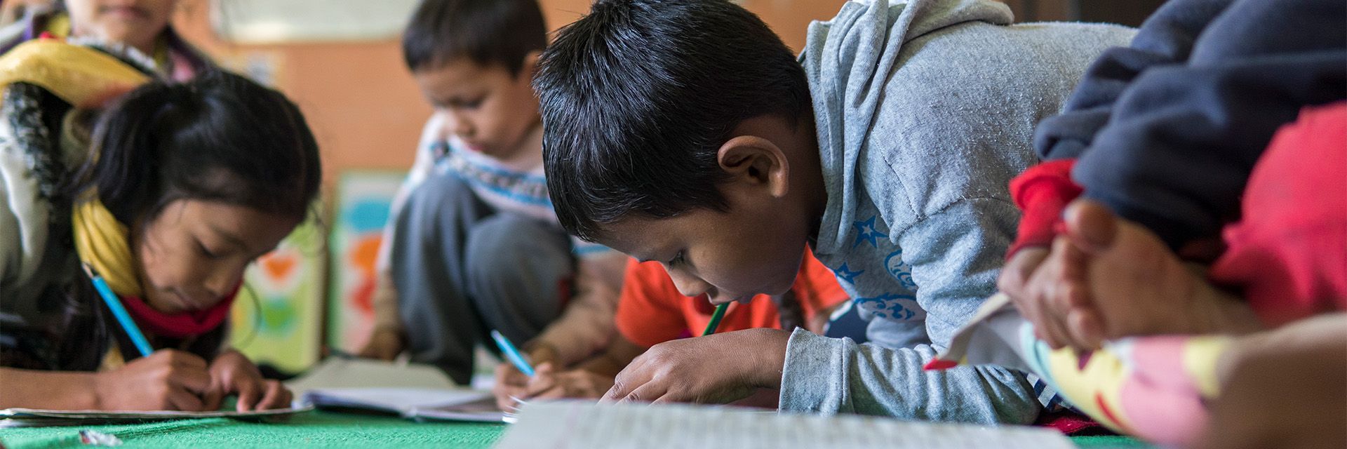 An image of children sitting on the ground in a classroom, writing in workbooks. 