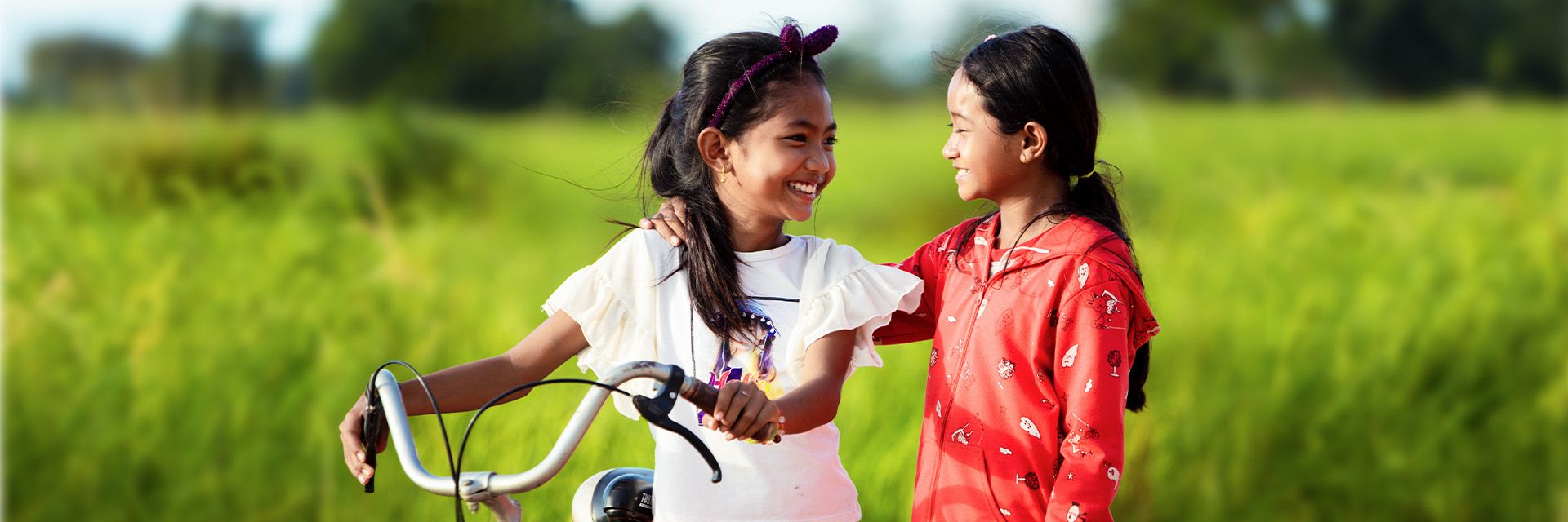 Two young girls smile at each other as they stand in a grass field with a bicycle.
