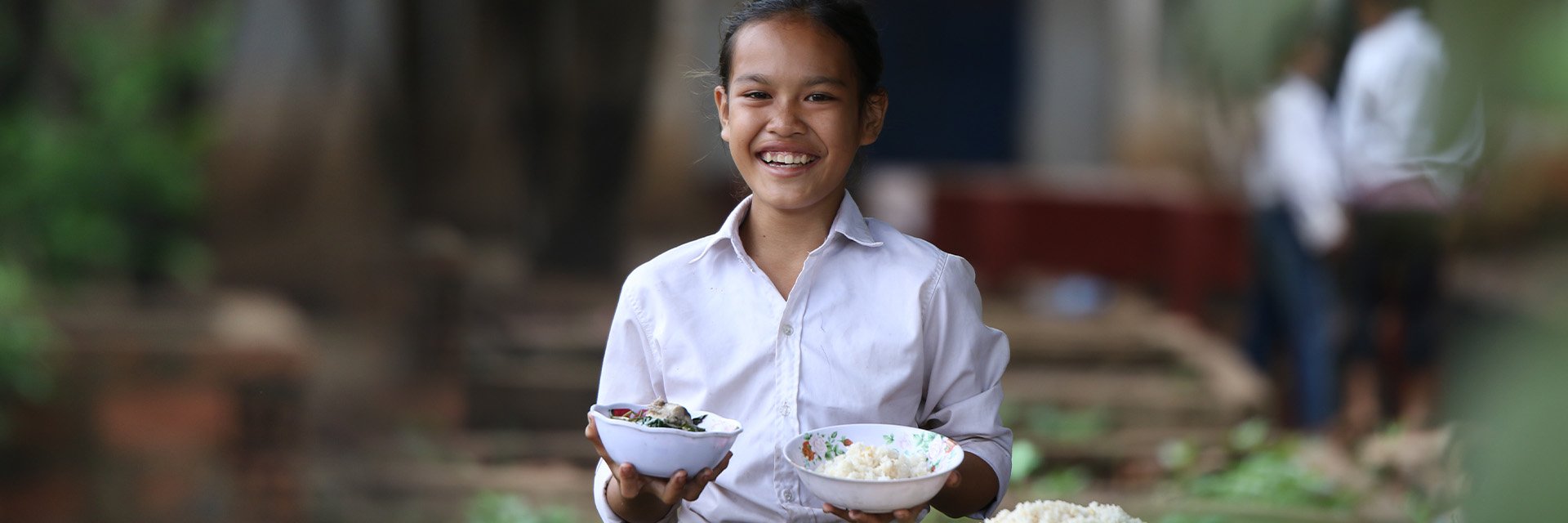 A young girl smiles widely while holding two white bowls of food. 