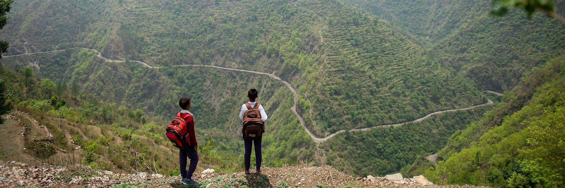 A wide-shot image of two people wearing backpacks, looking up at a massive green mountain area. 