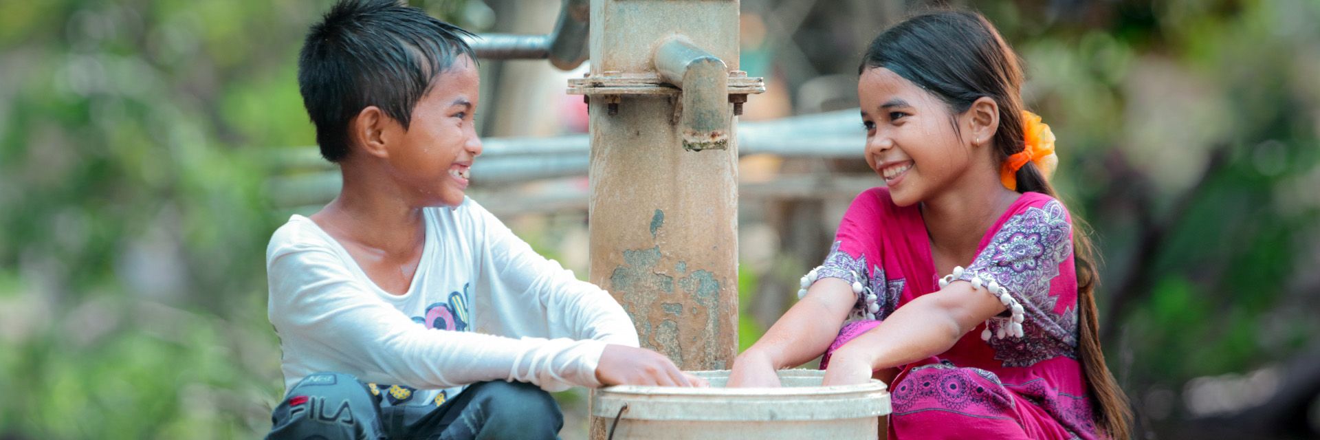 A young boy and girl smiling as they collect water from an outdoor faucet with a bucket. 