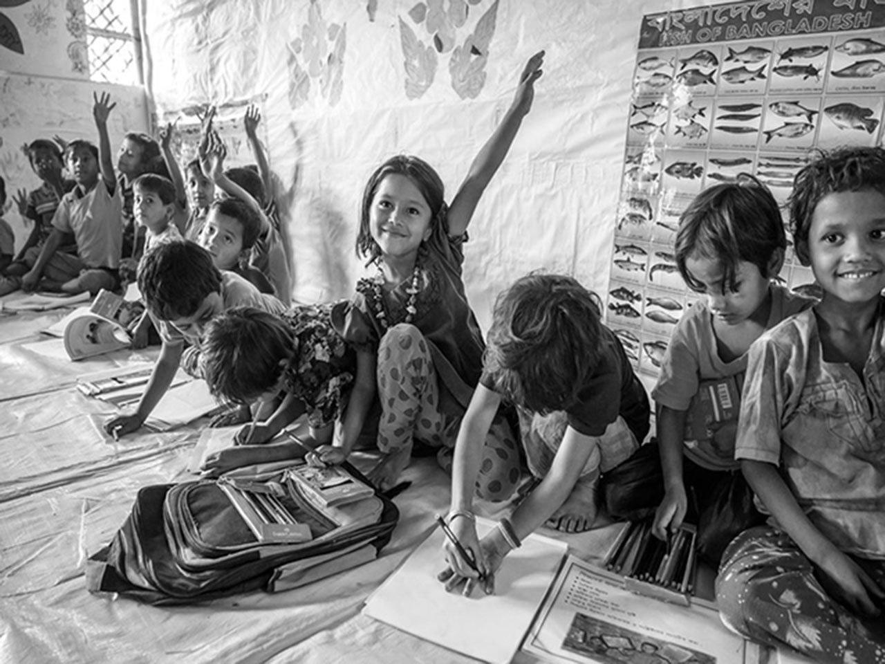Young children are working on a school activity while sitting on the floor. Some children are raising their hands and others are writing in their notebooks.