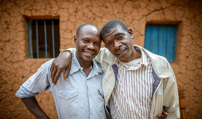 Two Rwandan men stand with their arms around eachother and a mud building behind them