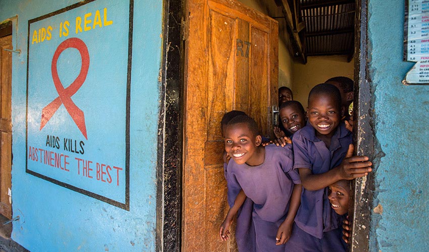 A group of kids in purple uniforms stand in a classroom doorway smiling, a nearby poster says “AIDS kills; abstinence is the best.”