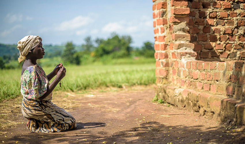 In Uganda, a young woman sits on the dirt ground outside a church and prays holding a cross.