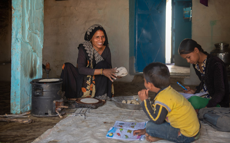 An Indian woman smiles at 2 children. They sit on the floor inside next to a small stove.