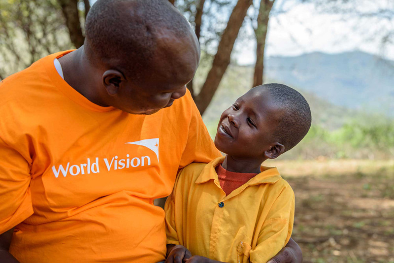 An adult male wearing an orange World Vision shirt gazes lovingly at a smiling little girl in West Pokot, Kenya.