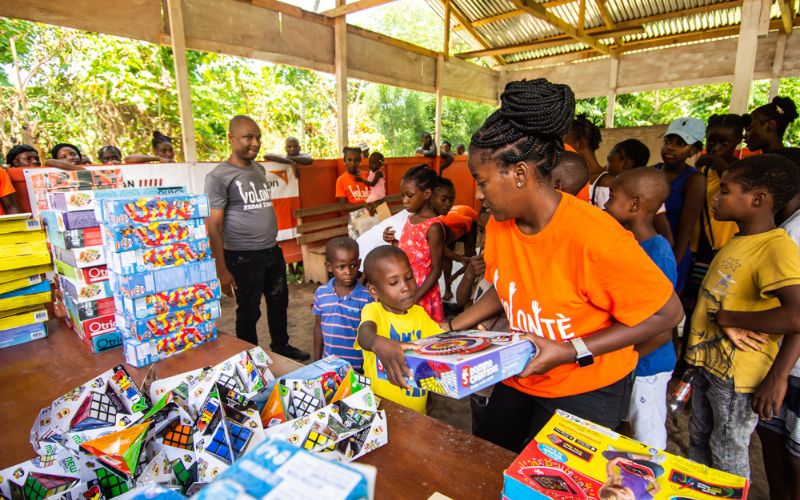A volunteer hands a box of toys to a child from a table filled with games and toys in front of her.
