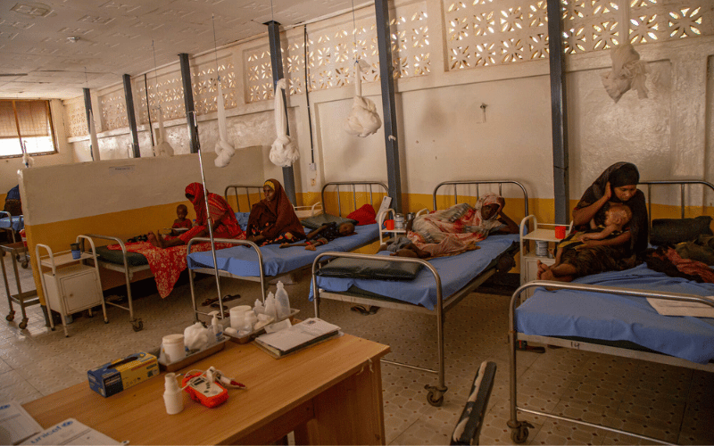 Four Somali women with their children sit and lie down on beds in a health clinic.