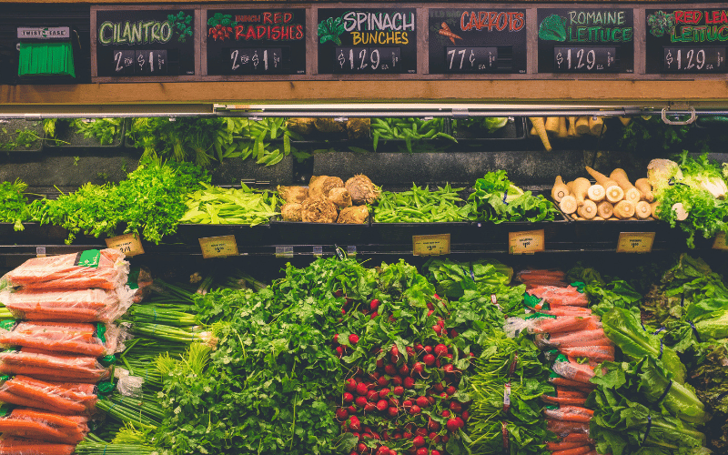 Rows of fresh vegetables in a grocery store sit under colourful signs with prices.