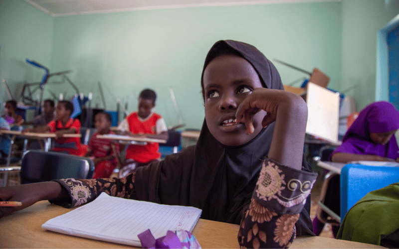 A young Somali girl in a hijab in her classroom sitting at her desk with a pencil and a notebook.