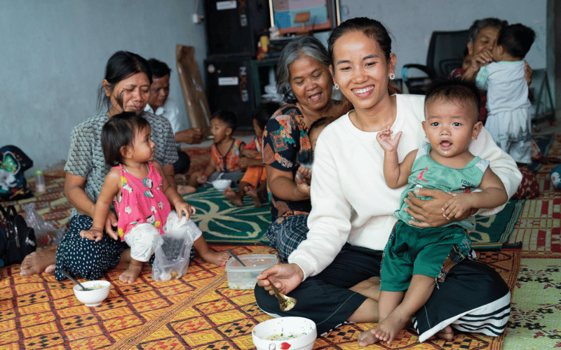 A group of Cambodian women sit on mats with bowls of food and their babies in their laps.