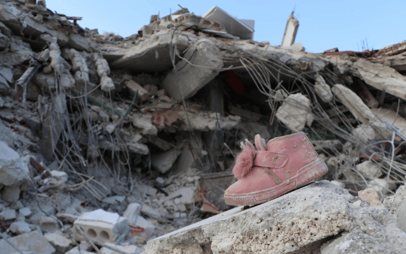 A pink shoe rests on top of a slab of broken concrete, amidst the debris of a building in the background.