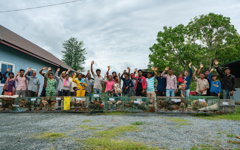 A crowd of farmers and their families stand behind crates of chickens, smiling and waving.