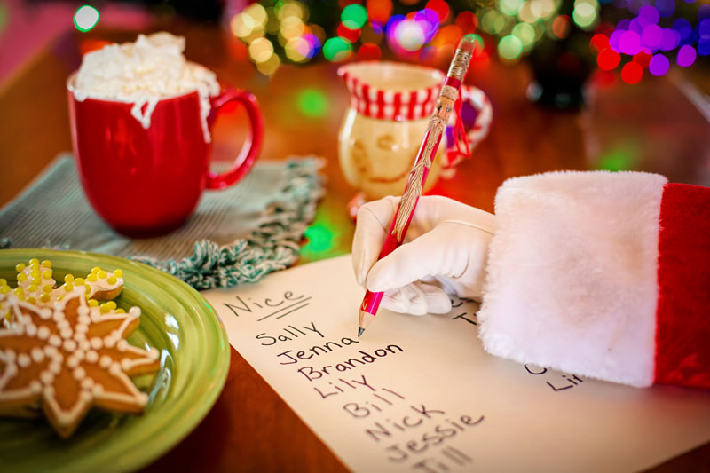 Santa’s hand writes names on his “Nice” list. Nearby is a green plate of cookies and a red mug topped with whipped cream.