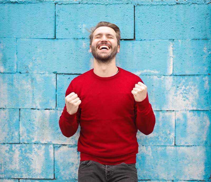 A man in a red sweater and black jeans has a happy expression on his face as he stands in front of a blue wall.