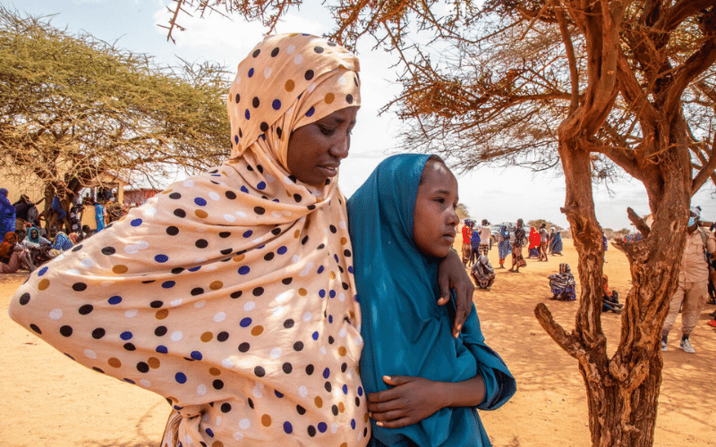 A woman and girl stand close together outdoors, in a camp for people in Somalia displaced due to threats like climate change.