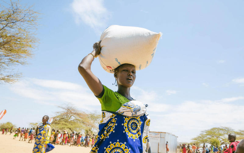 A woman walks carrying a bag of grain on her head.